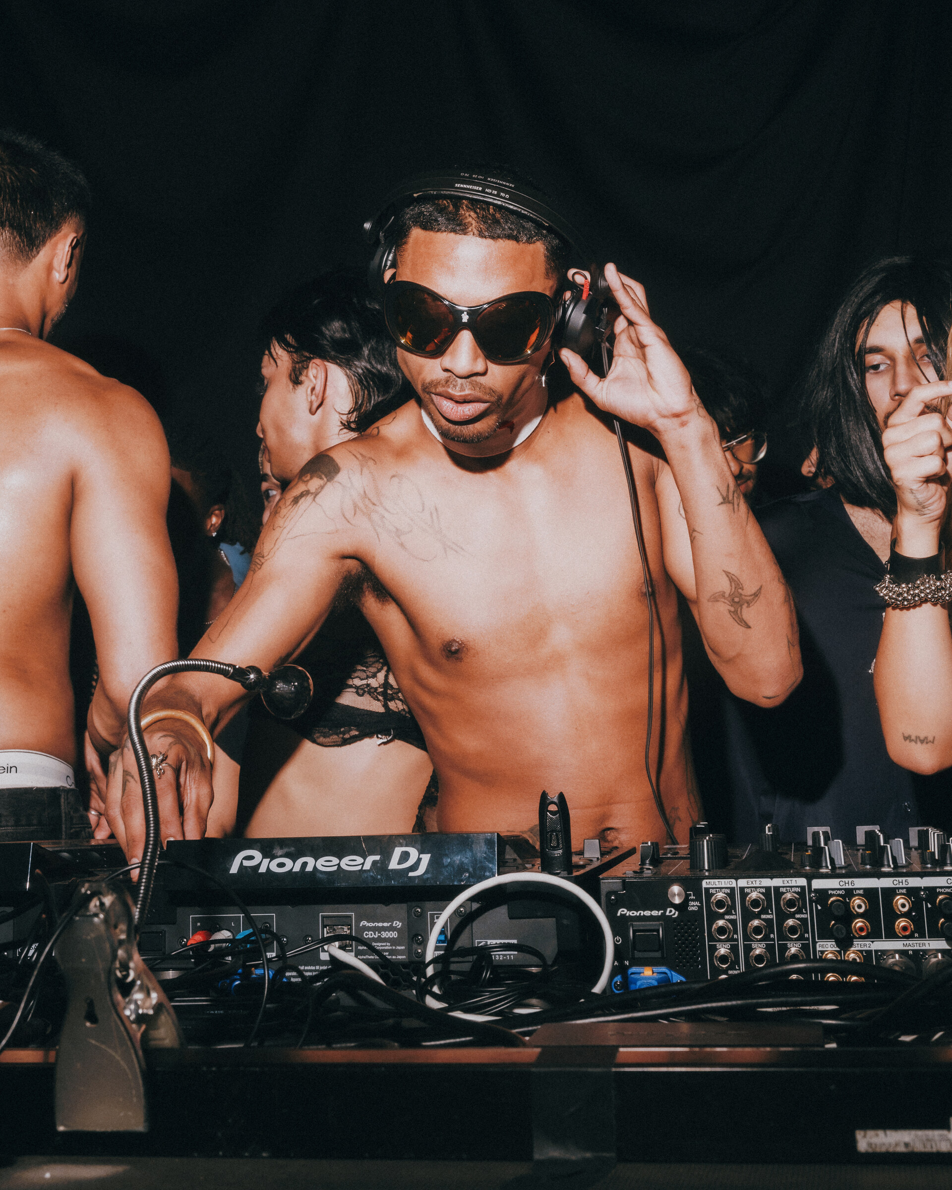 Shirtless DJ wearing sunglasses and headphones behind Pioneer DJ decks at Pep Rally Pride 2026